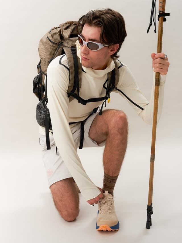 A man in hiking gear, sunglasses, and backpack kneels, tying his shoe and holding a trekking pole.