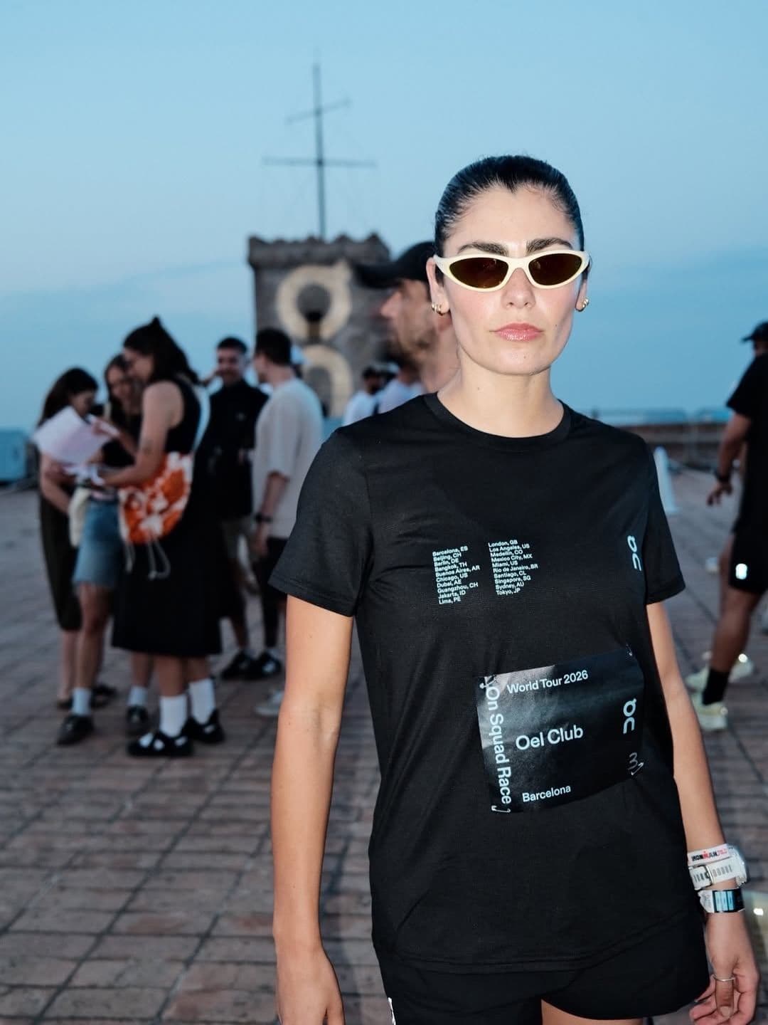 A woman in a black t-shirt with "Oel Club World Tour 2026 On Squad Race" text and white sunglasses looks at the camera, with blurred people behind her outdoors.