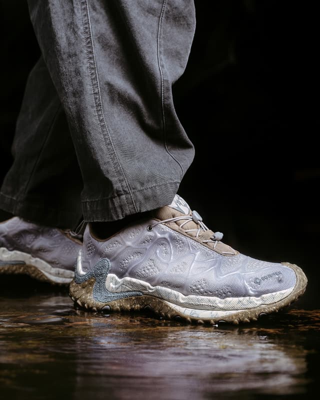 Close-up of a person wearing dark grey pants and light grey, textured GORE-TEX sneakers with a rugged sole, standing on a wet, reflective surface.