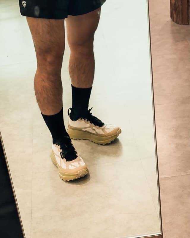 A person's legs wearing black shorts, socks, and cream-colored sneakers with black laces, reflected in a mirror.