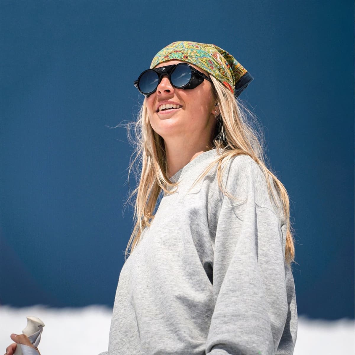 Smiling woman wearing a bandana and glacier glasses, holding a ski pole against a blue sky and snow.