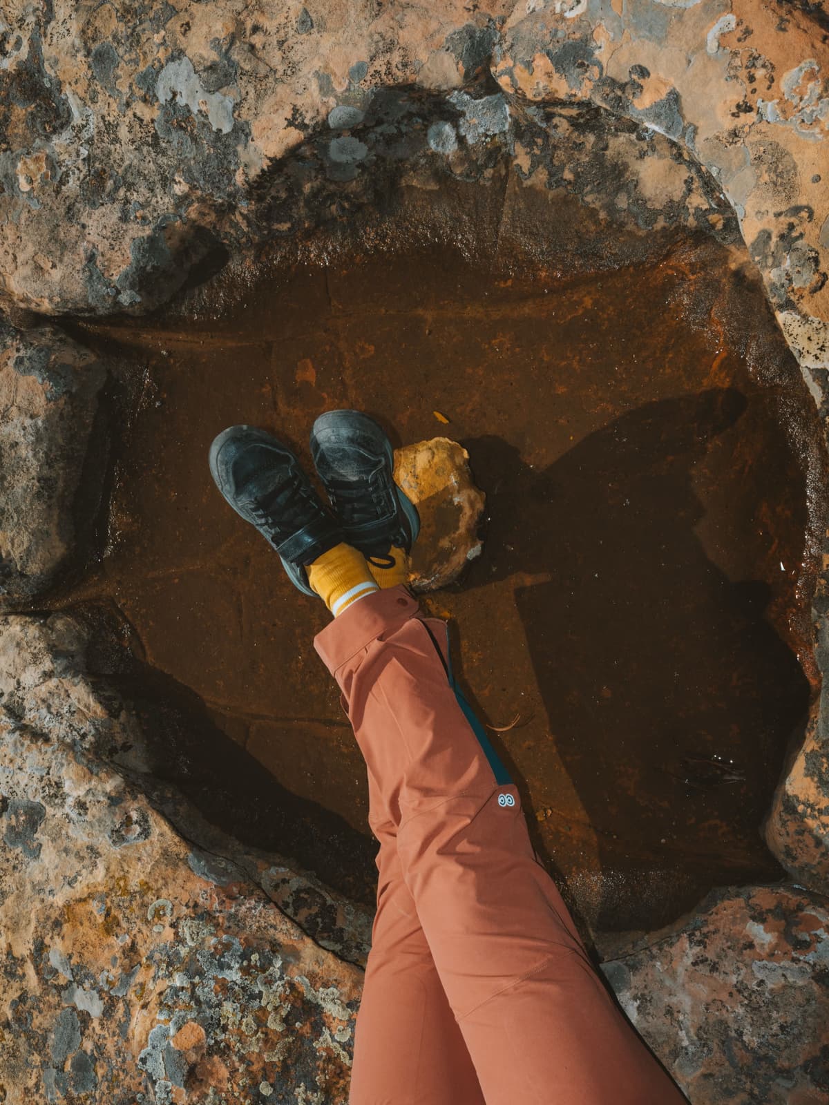 A person's feet in black hiking boots and orange pants resting in a rock pool.