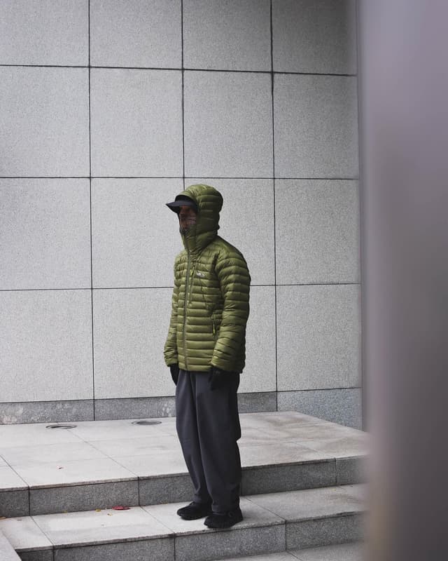 A person in a green hooded puffer jacket, black cap, and grey pants stands on stone steps against a tiled wall.