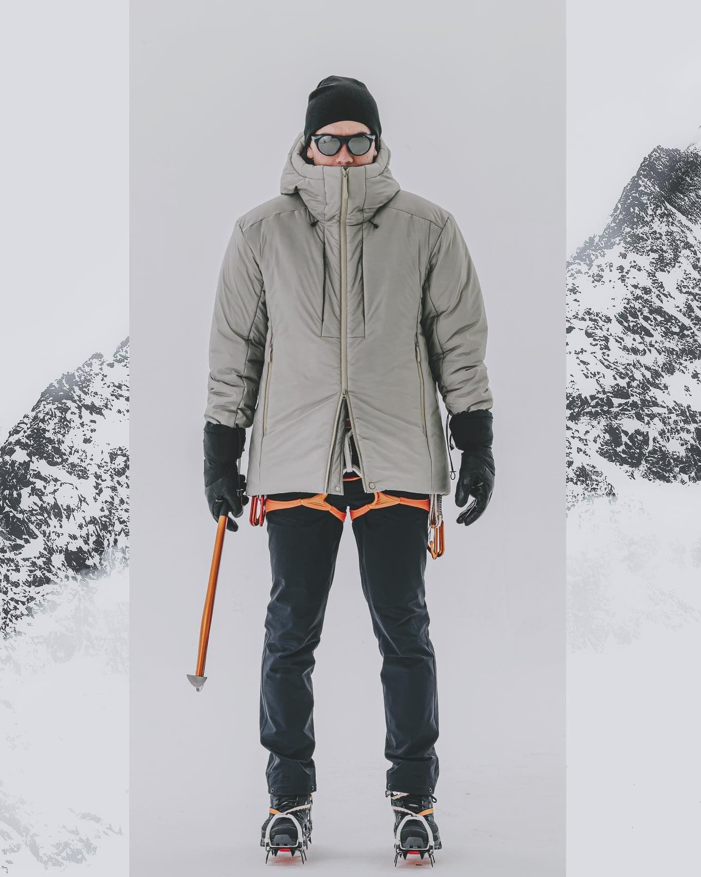 A person in a grey winter jacket, beanie, and sunglasses holds an ice axe, wearing a harness and crampons. Snowy mountains flank a plain background.