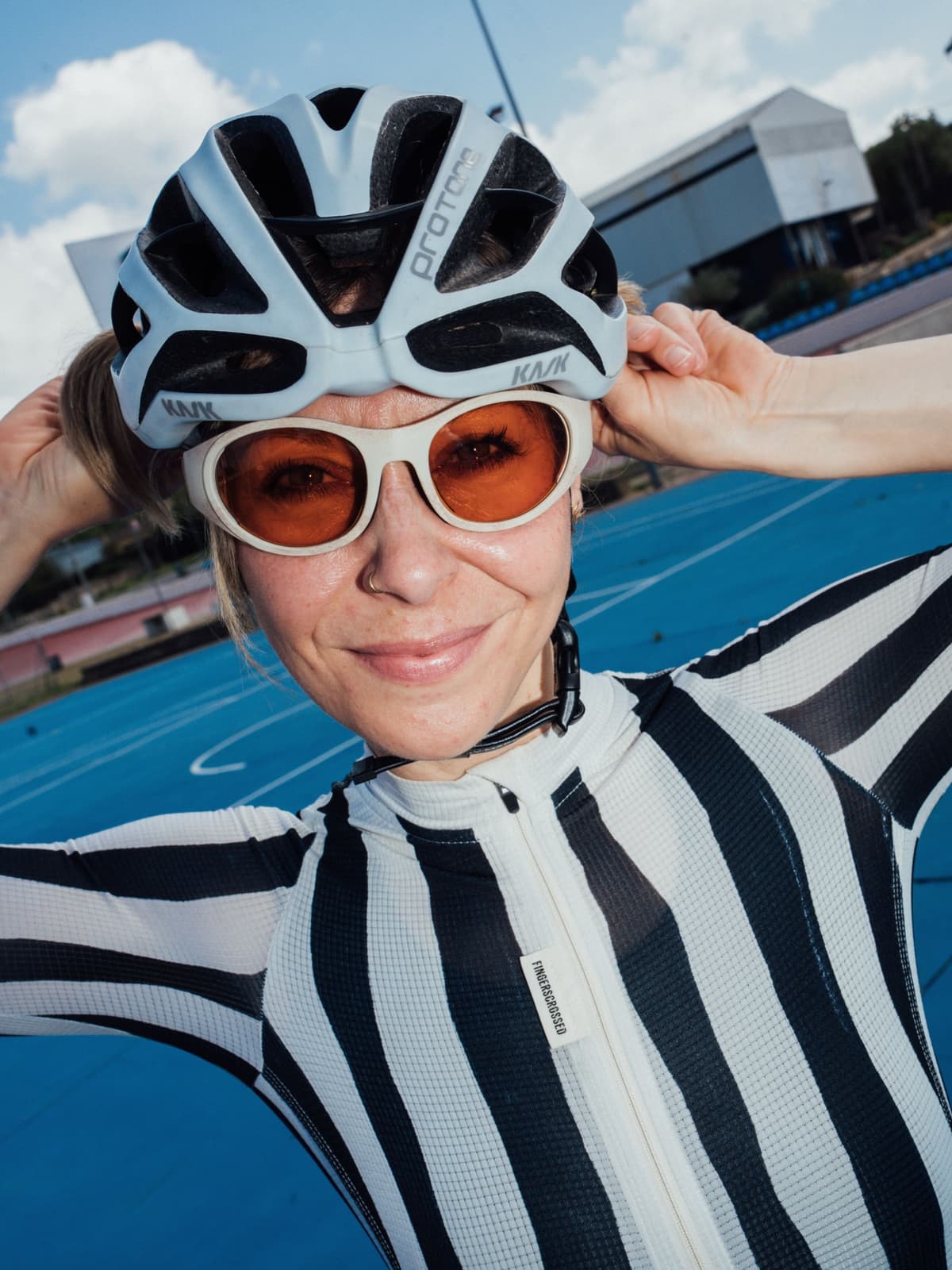 A woman in a blue cycling helmet, orange sunglasses, and a black and white striped jersey smiles at the camera on a blue track.