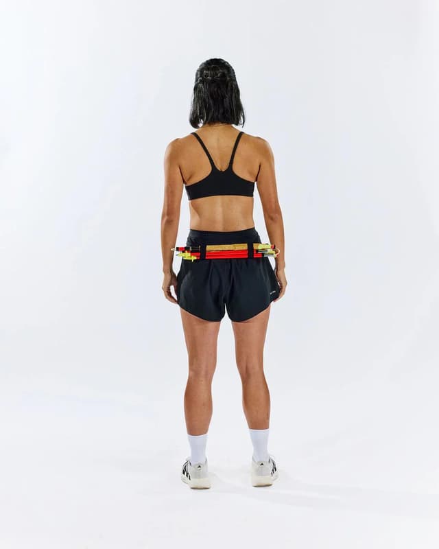 A person from behind wears a black sports bra and shorts, with colorful batons secured by a belt around their waist.