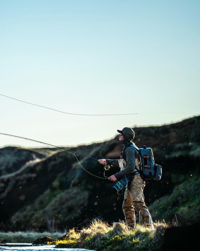 An angler in waders casts a fly fishing line on a sunlit river bank.