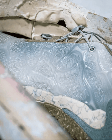 Close-up of a shoe with a light blue mesh upper, clear wavy overlays, grey laces, and a cream and blue speckled sole.