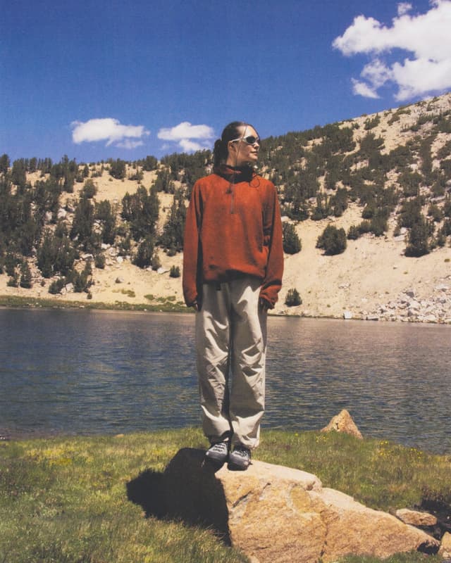 A person in a red fleece and sunglasses stands on a rock by a mountain lake, with a treed hillside under a blue sky.