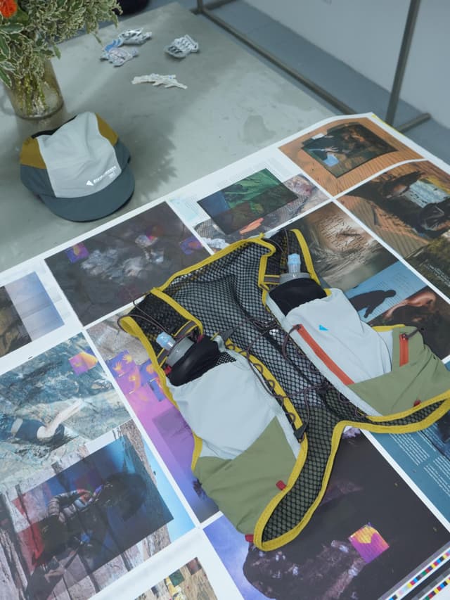 A grey and olive green running hydration vest with yellow trim and two water bottles lies on a table covered with printed images, with a matching cap on the floor.