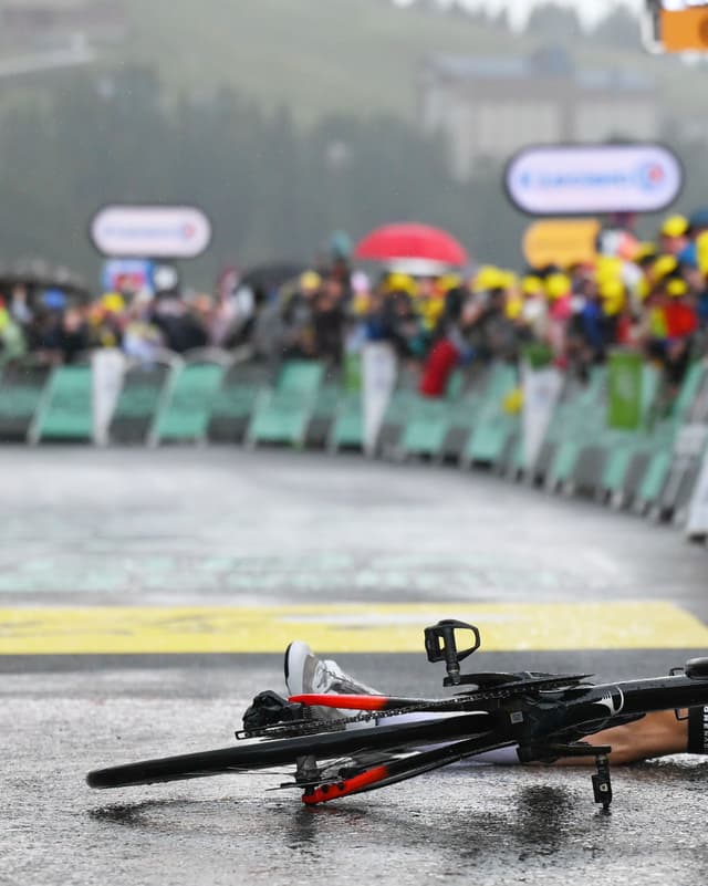 A black road bicycle lies on wet asphalt next to a person's leg, with blurred spectators and event barriers in the background.