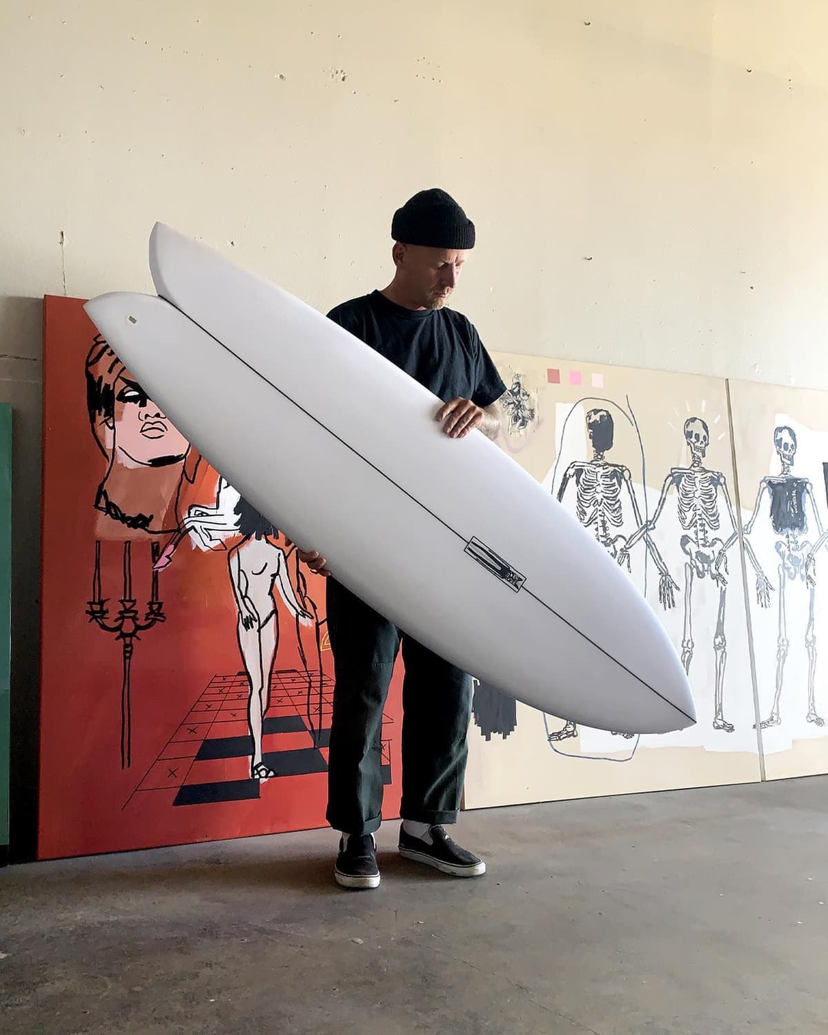 A man in a black beanie holds two white surfboards in front of large, graphic paintings.