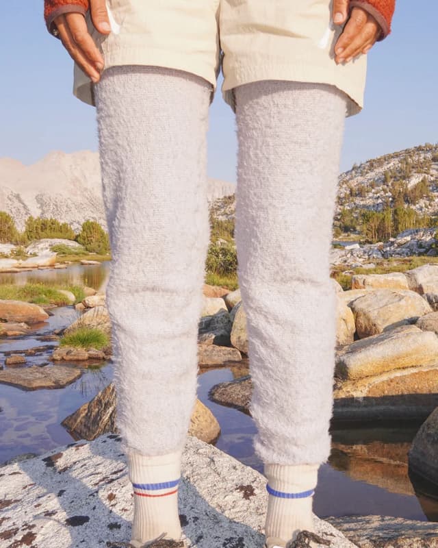 A person stands on rocks by a stream, wearing tan shorts over fluffy gray leg warmers and white socks with colored stripes. Mountains are in the background.
