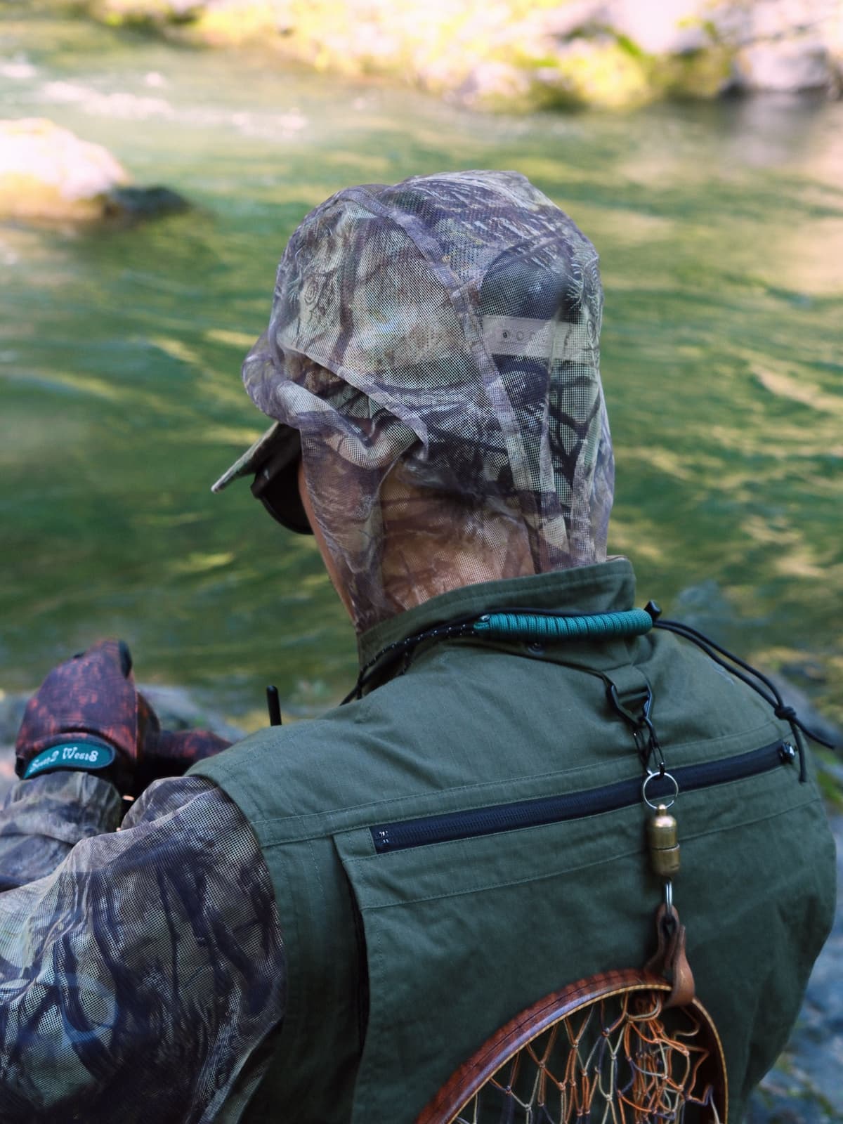 A person in camouflage and a green vest, with a fishing net on their back, looks out at a river.