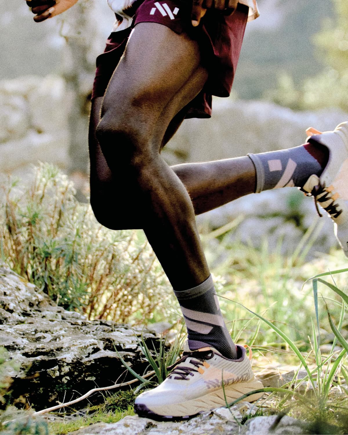 A runner's legs in maroon shorts, patterned socks, and trail shoes stride over a rocky path.