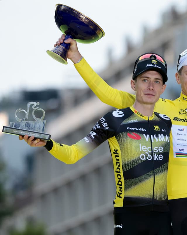 A cyclist in a black and yellow jersey holds a large blue trophy overhead and a smaller bicycle trophy in his other hand.