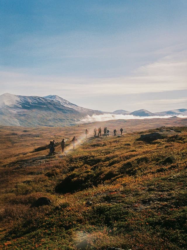 A group of hikers walks on a trail through a vast, autumnal landscape with snow-capped mountains in the distance.