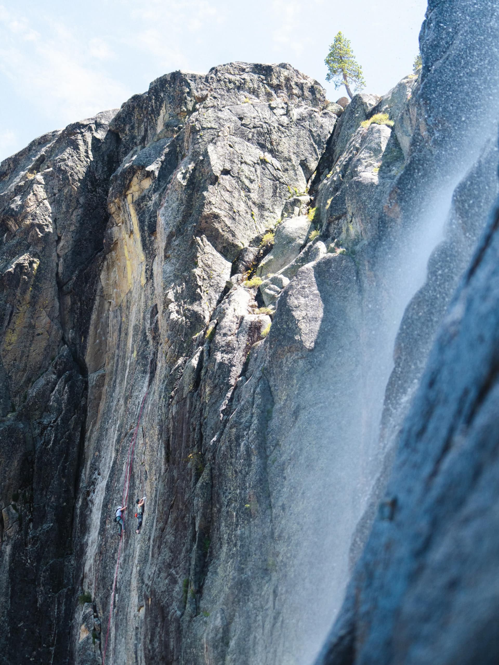 Two climbers hang from ropes on a sheer cliff face beside a rushing waterfall. A lone tree stands at the cliff's peak.