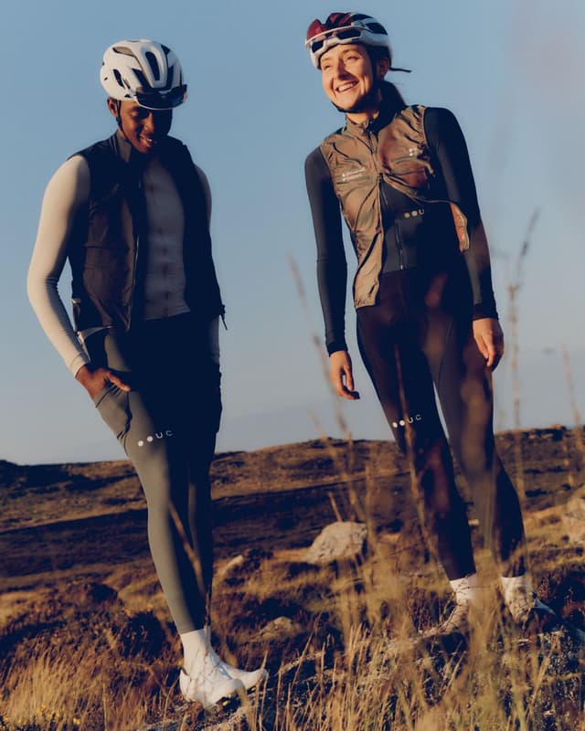 Two smiling cyclists, a man and a woman, wearing helmets and athletic vests, stand on a sunlit, rocky, grassy landscape.