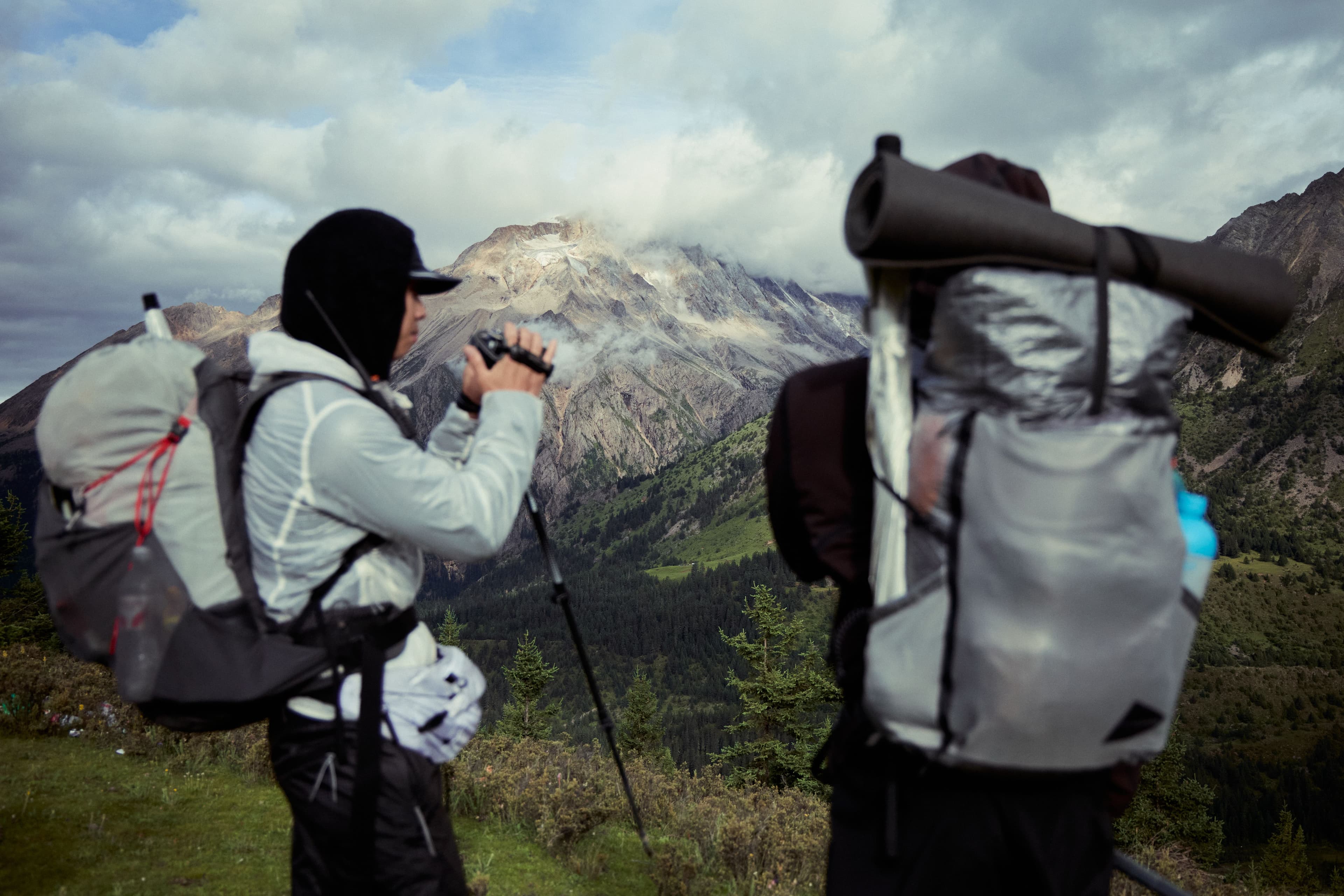 Two hikers with backpacks view a vast mountain landscape; one uses binoculars.