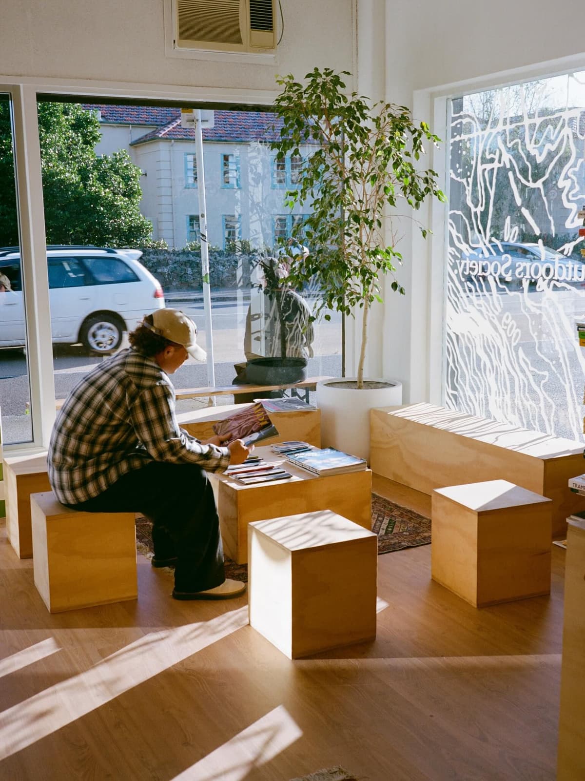 A person reads a magazine in a sunlit room furnished with simple wooden blocks, looking out a large window.