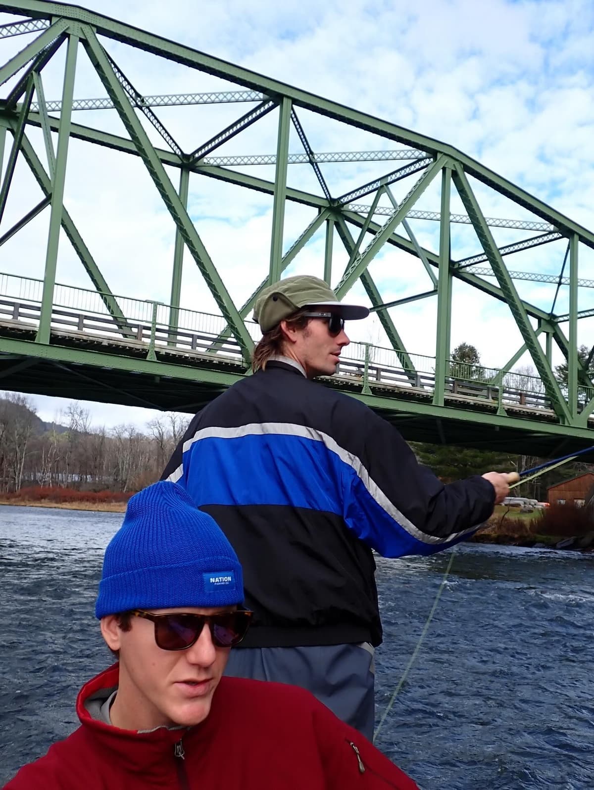 Two men on a river, one fly fishing, under a green truss bridge.
