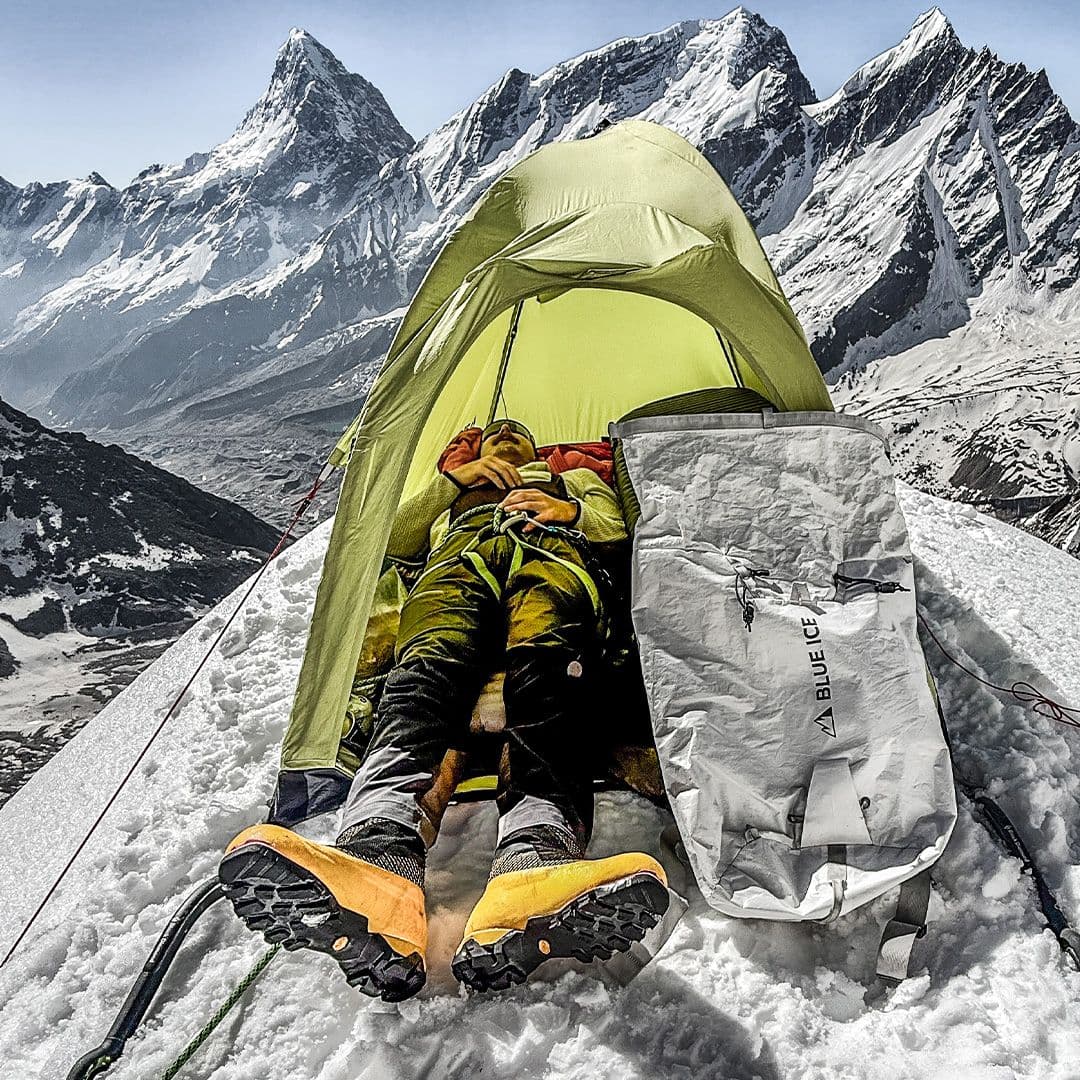 A climber rests in a green tent on a snowy mountain, with towering peaks in the background.