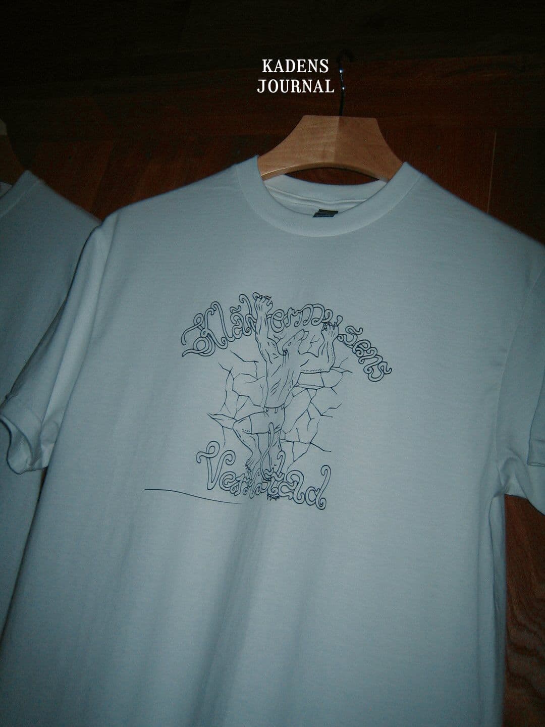 Light-colored t-shirt with a dark line art graphic depicting the words "Illumination" and "Verdad" and an abstract figure breaking through a cracked design, hanging on a wooden hanger.