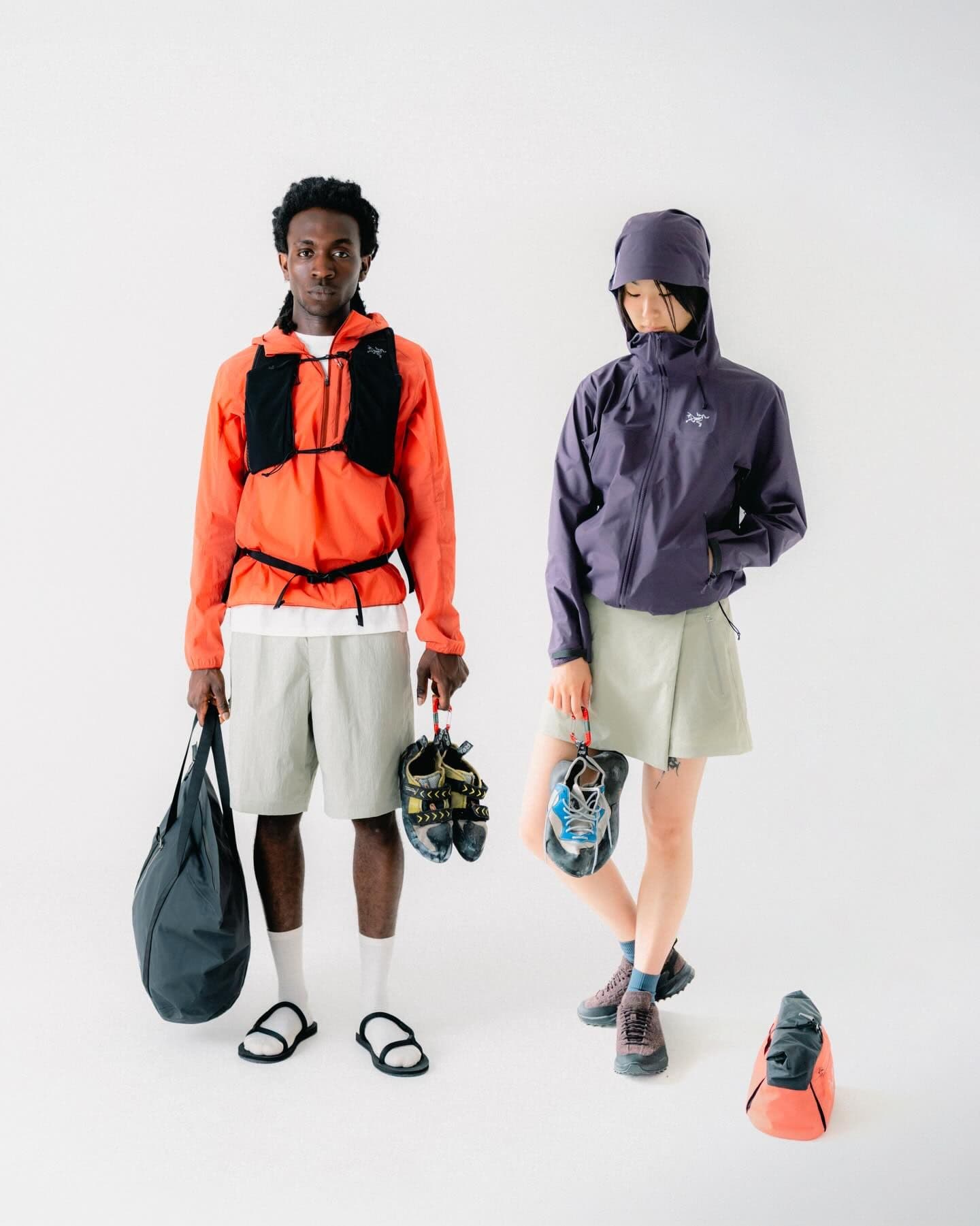 Two people in Arc'teryx outdoor gear. A man in an orange jacket, shorts, and sandals holds a black bag and climbing shoes. A woman in a purple jacket, skirt, and hiking shoes holds climbing shoes.