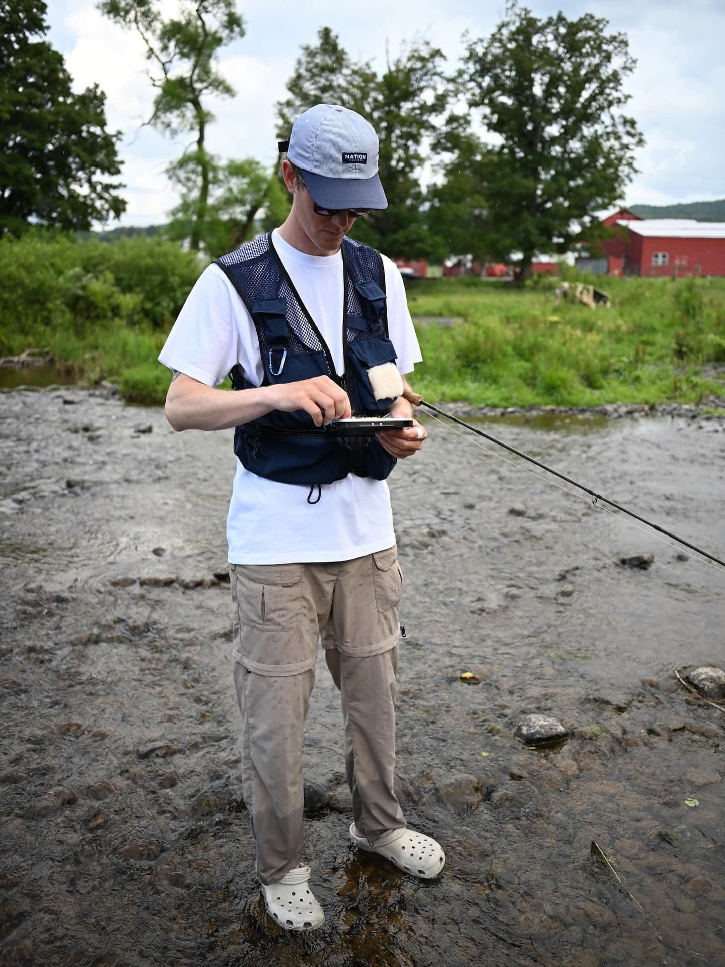 A young man in a fishing vest and hat stands in a shallow river, adjusting his fishing rod.