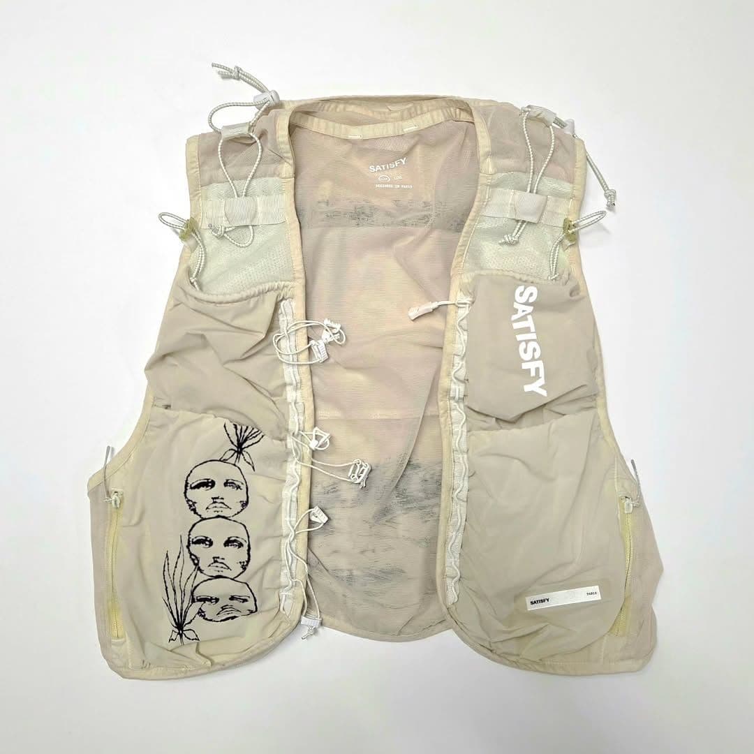 A cream-colored running vest with multiple pockets, a "SATISFY" logo, and a graphic of three stacked faces.