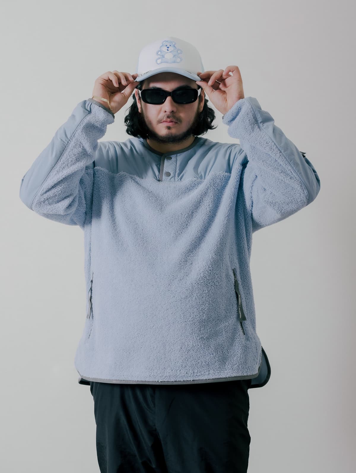 A man in a light blue fleece pullover, black sunglasses, and a white cap with a blue teddy bear, adjusting his hat.