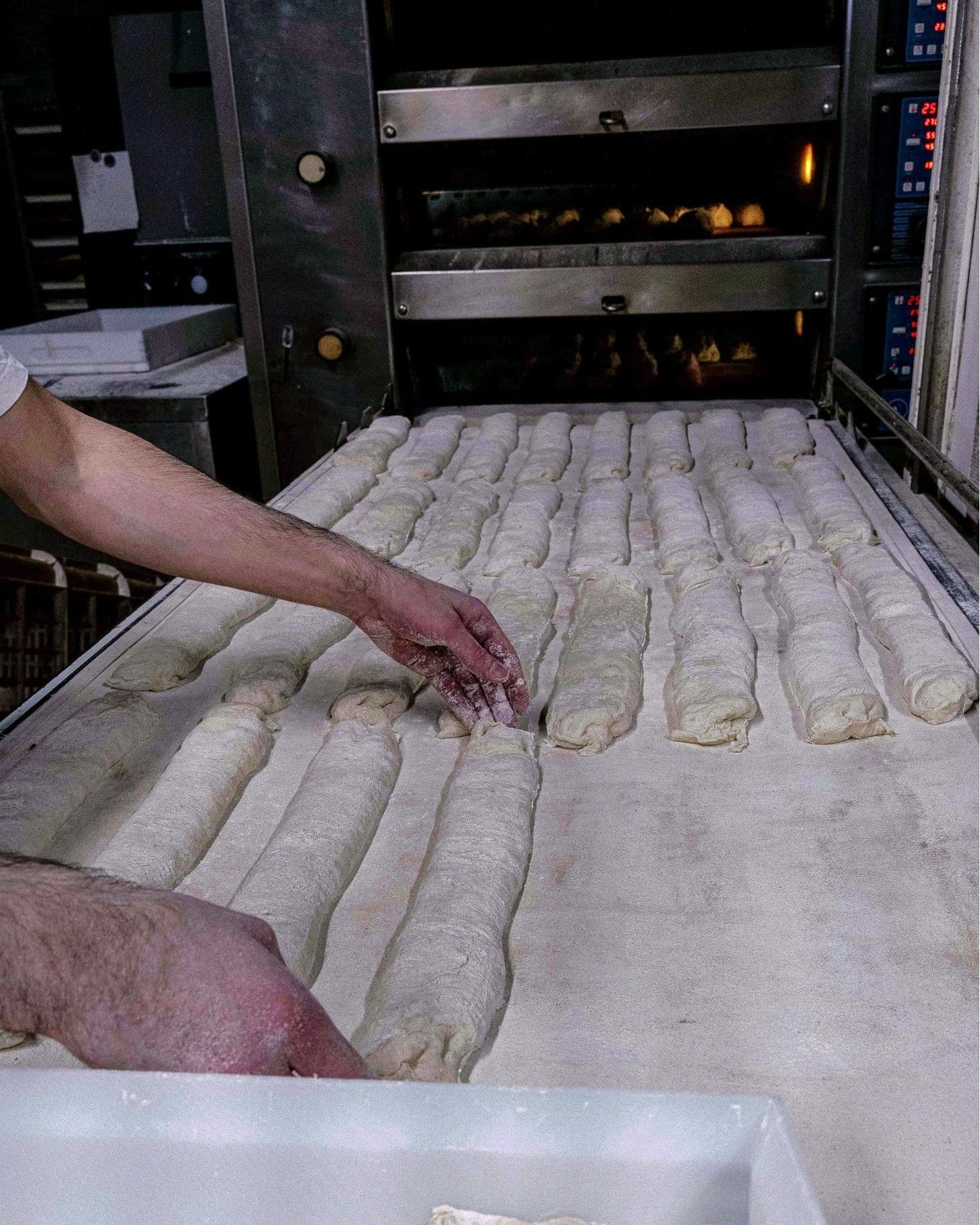 A baker arranges rows of unbaked baguette dough on a floured surface in front of an oven with baking loaves.