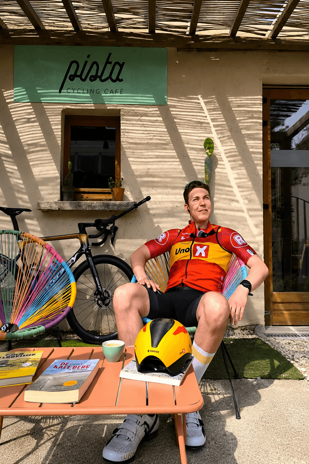 A male cyclist in a red and yellow jersey relaxes in a chair outside "Pista Cycling Cafe," next to his bike, with his helmet and coffee on a table in dappled sunlight.