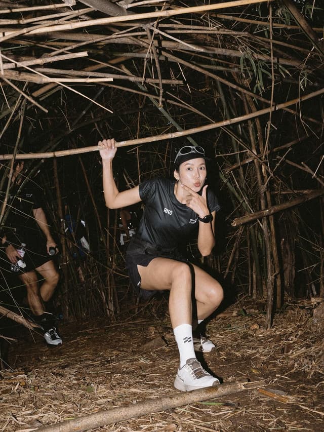 A woman in athletic gear playfully poses while holding a bamboo pole in a dark bamboo forest.