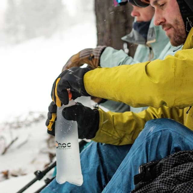 A person in a yellow jacket and black gloves holds a transparent CnOC Outdoors flexible water bottle in a snowy outdoor setting.