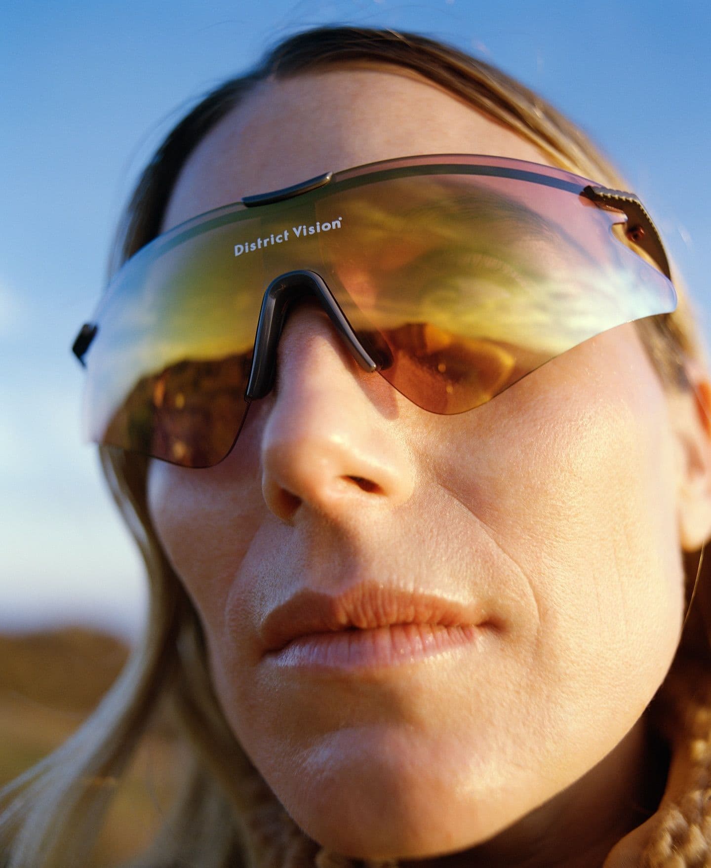 Close-up of a woman wearing District Vision sunglasses with amber reflective lenses, showing a landscape reflection, against a blue sky.