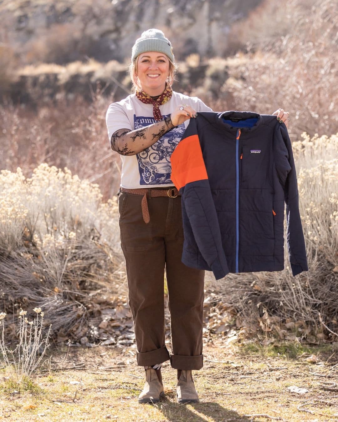 A smiling woman outdoors holds up a dark blue Patagonia jacket with an orange shoulder panel.