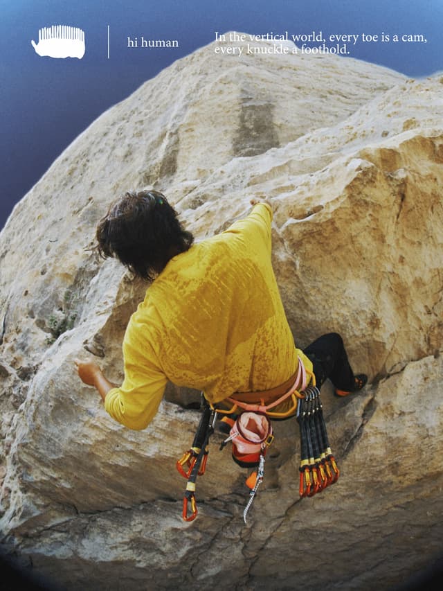 A person in a yellow shirt and black pants climbs a rock face, seen from behind, wearing a climbing harness with gear.