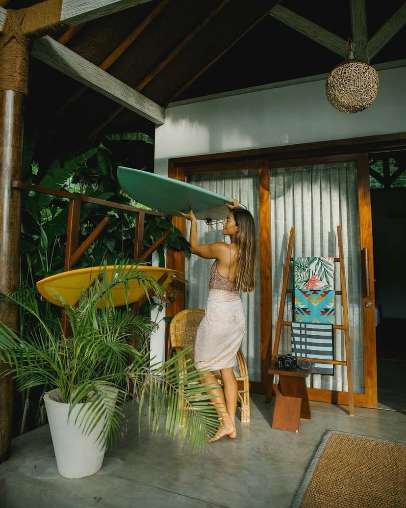 A woman holds a light blue surfboard on a tropical patio with lush plants.