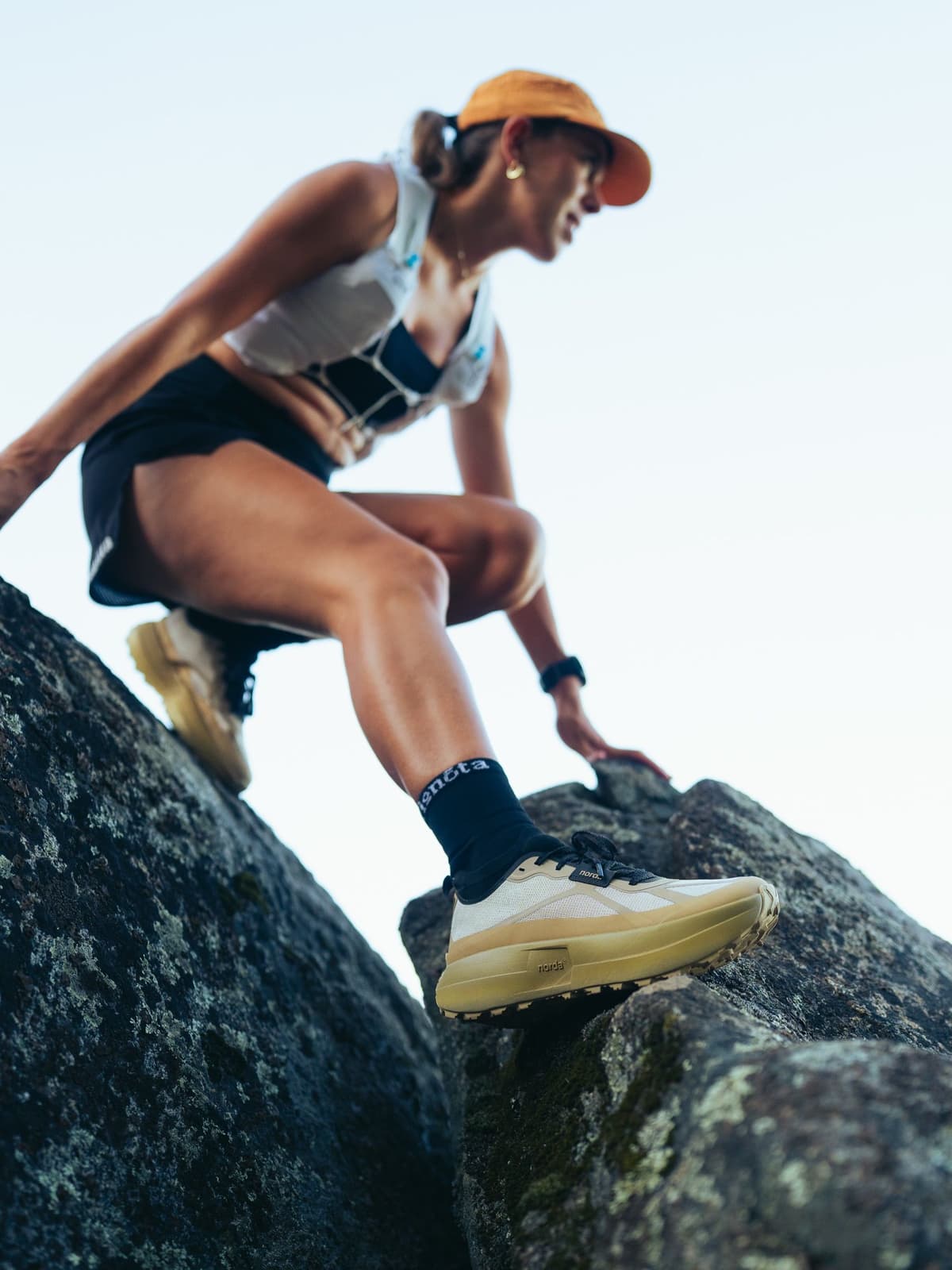 A woman in an orange hat, white vest, and trail running shoes climbs over dark rocks.