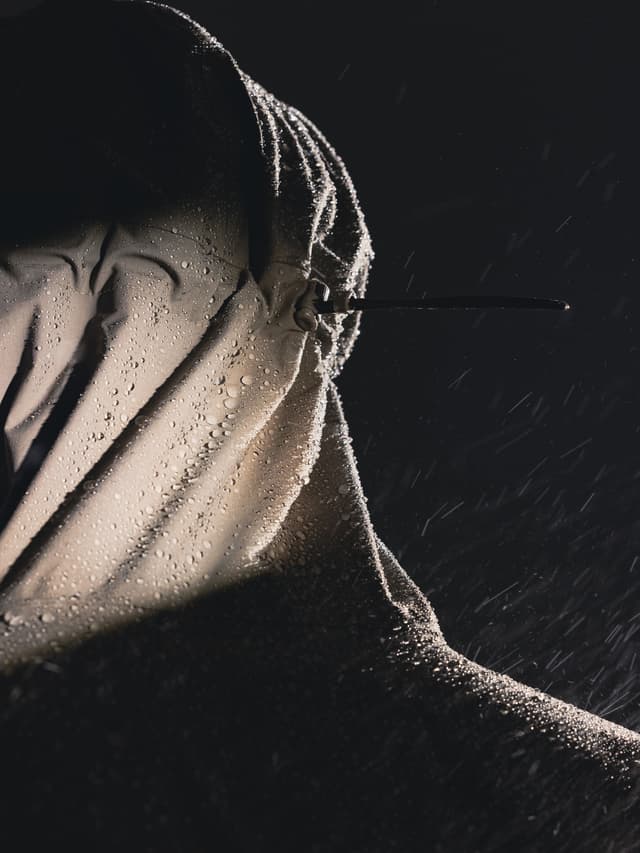 Close-up of a light-colored, waterproof hooded jacket covered in rain, against a dark background.