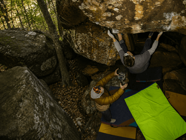 A boulderer hangs upside down from a rock overhang while a spotter below takes a photo, surrounded by crash pads in a forest.