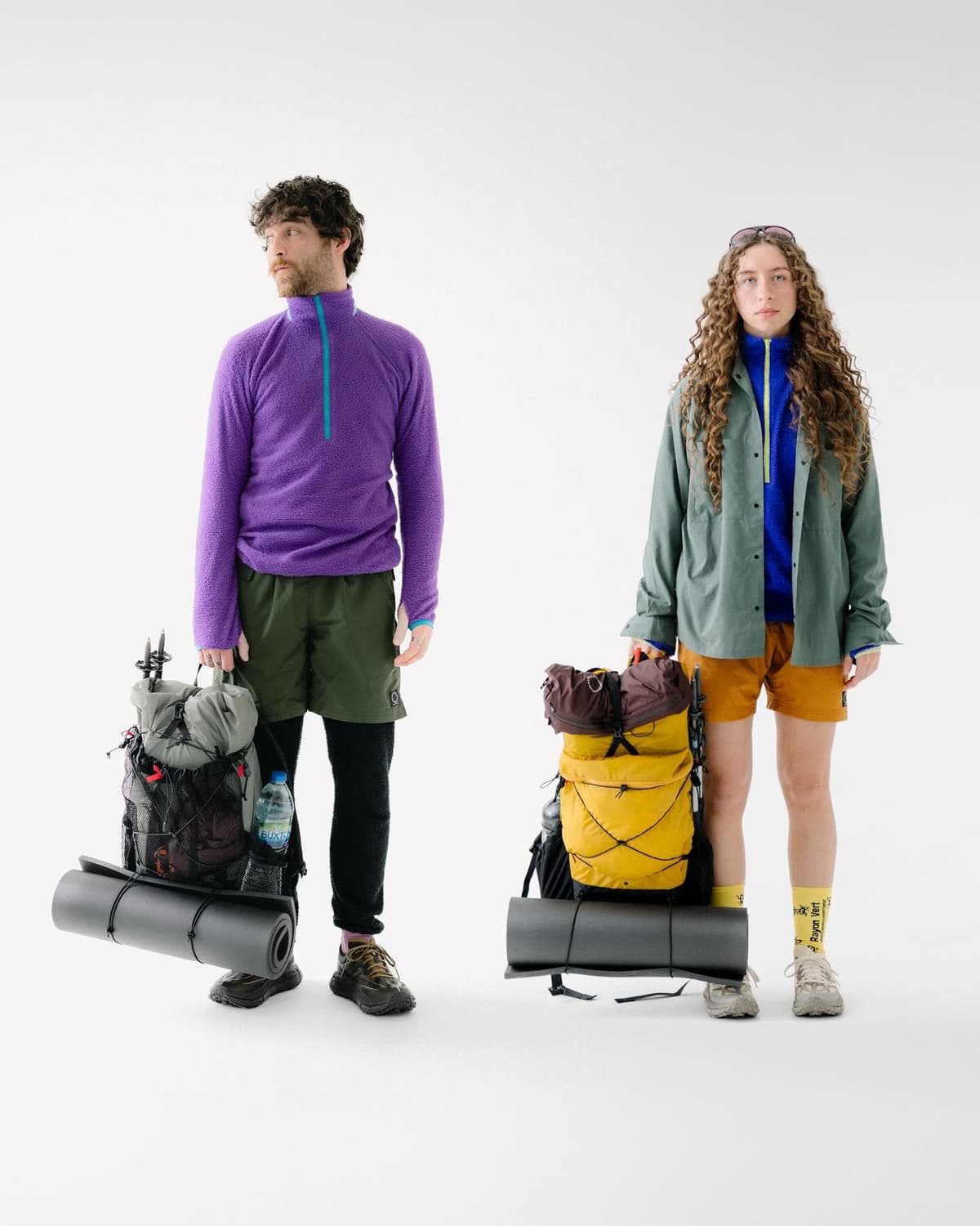 A man and woman in outdoor clothing carry large backpacks with sleeping pads against a white background.