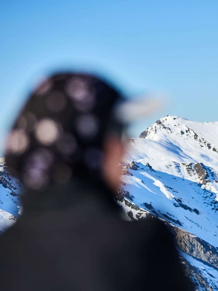 Out-of-focus person in a patterned hat looking at sharp snow-capped mountains under a blue sky.
