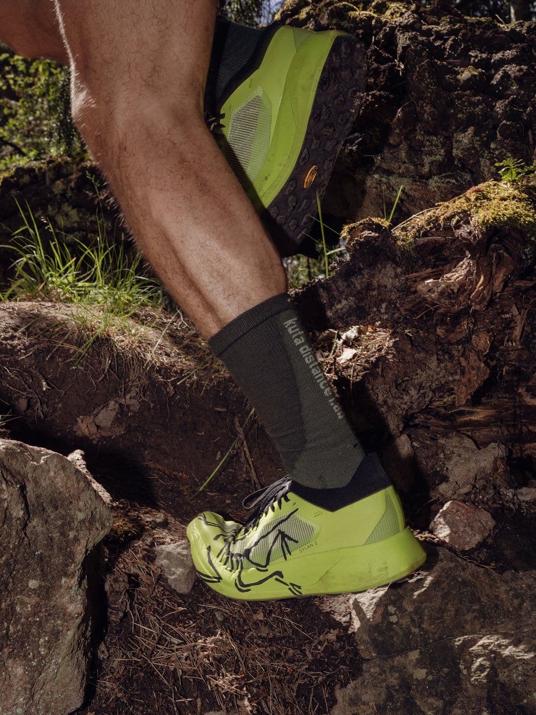 A person's legs in bright lime green trail running shoes and dark socks step on rocks and roots on a trail.