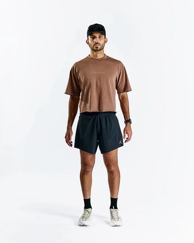 A man wearing a brown t-shirt, black shorts, black cap, and sneakers stands on a white background.