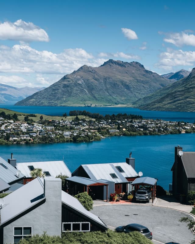 Modern houses on a hillside overlooking a town by a blue lake with large mountains in the background.