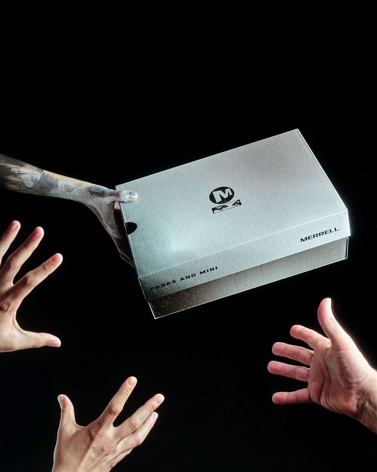 A hand with a rough, dark texture holds a silver Merrell shoebox above two pairs of hands reaching up from below, against a black background.
