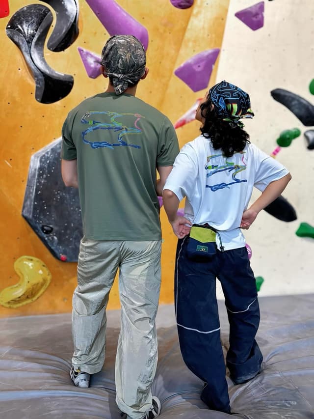 Two people from behind, wearing bandanas and matching graphic tees, stand on a mat in front of a climbing wall.
